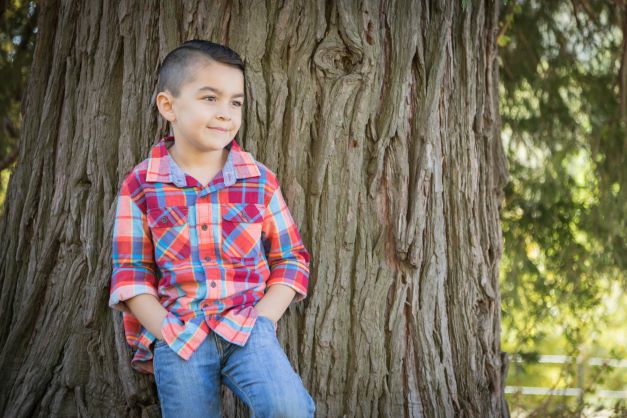 Adorable little boy in plaid shirt and jeans leaning against tree