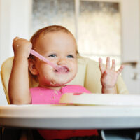 baby feeding herself cereal with spoon in highchair