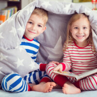 two kids reading a book in their pj\'s under a blanket