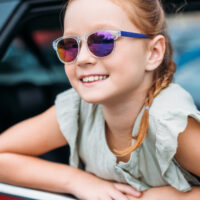 little girl wearing kids sunglasses leaning out of car window and smiling