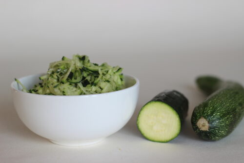 shredded zucchini in a bowl next to a halved zucchini