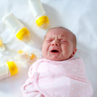 crying newborn baby girl laying on bed surrounded by bottles and pacifier