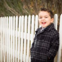 Little boy in plaid coat standing by white fence