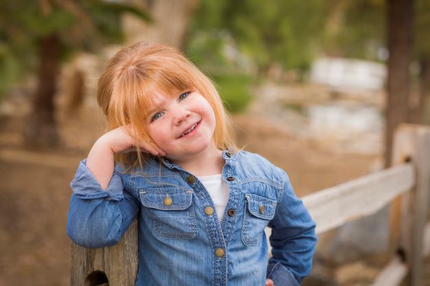 Little girl with red hair posing at the park against a fence
