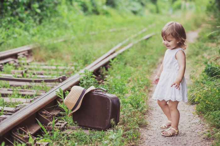 Toddler girl in sandals with suitcase by train track